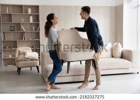 Happy married couple enjoying moving into new house. Excited homeowners, renters, man and woman carrying furniture, holding armchair, talking, joking, laughing, feeling joy. Relocation concept Royalty-Free Stock Photo #2182327275