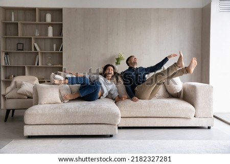 Funny excited millennial spouses of new homeowners falling on big soft couch at home. Young couple celebrating moving into first common house. Real estate, renovation, furniture for hoe concept Royalty-Free Stock Photo #2182327281