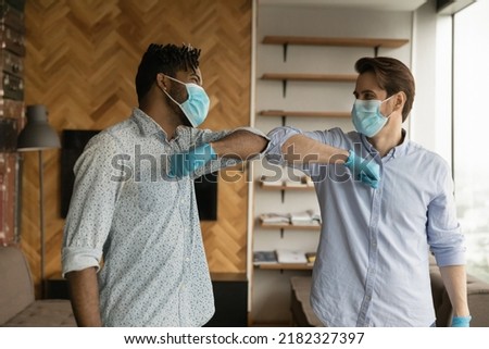 Happy diverse multiethnic male colleagues in facial masks rubber gloves greeting in office during covid-19. Smiling international men in medical protective gear touch elbows. Corona virus concept. Royalty-Free Stock Photo #2182327397