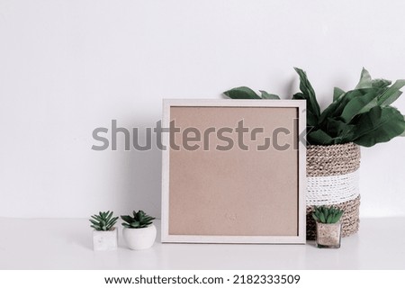 Blank wooden square frame and green houseplants flowers on table against white wall background. Mockup Template for your design, text.