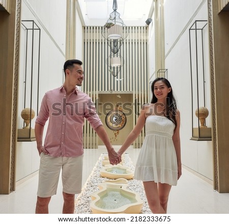 Young asian couple walking through luxury spa or beauty salon. Weekend relaxation, celebrating honeymoon or anniversary. Man and woman hold hands and posing against modern arabic design background
