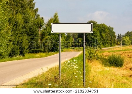 Asphalt road with white empty sign on roadside at city entrance against sunny landscape background. 