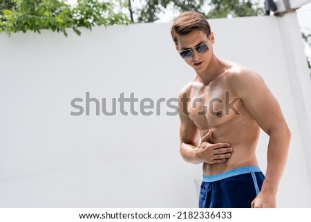man in sunglasses and swimming trunks applying sunscreen on muscular torso Royalty-Free Stock Photo #2182336433