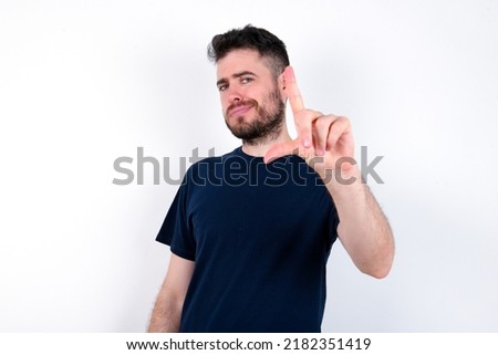 Young caucasian man wearing black T-shirt over white background making fun of people with fingers on forehead doing loser gesture mocking and insulting.