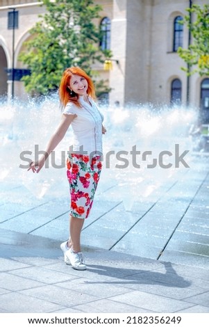 Summer concept. Happy red-haired Caucasian girl in a white blouse and midi skirt with rose print jumping for joy at the fountain. Water splash background on a hot summer day