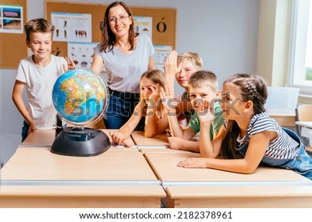Cute happy schoolchildren with teacher looking at camera raising hands, get ready to answer at geography lesson with globe. Friendship, interaction teamwork concept. Royalty-Free Stock Photo #2182378961