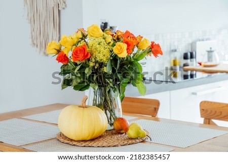 A vase of yellow and orange rose flowers, fresh pumpkin, apple and pear on a kitchen table counter with white modern kitchen background. Autumn home decor for Thanksgiving and Halloween holiday