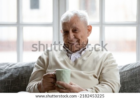 Cozy winter evening. Portrait of happy elderly man resting on soft couch at home enjoying cup of hot blended tea. Grey haired retired male looking at camera with smile drinking aromatic natural coffee Royalty-Free Stock Photo #2182453225