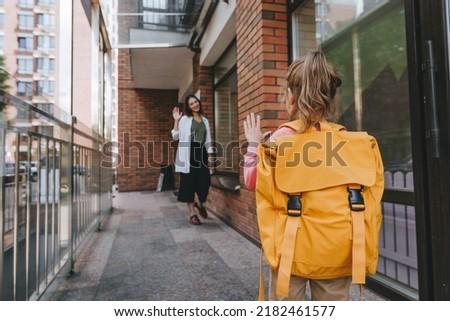 Little girl with backpack waving her mother saying goodbye near the school. Back view. Back to school concept. Royalty-Free Stock Photo #2182461577