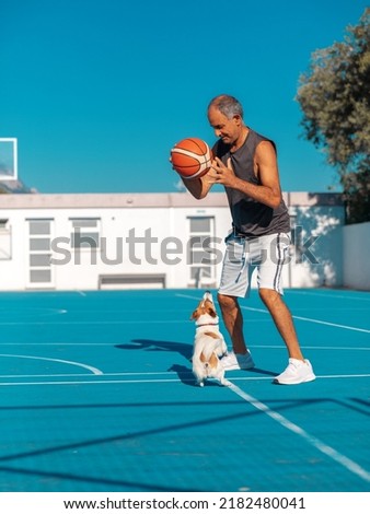 Turkish Cypriot elderly athletic man having fun playing basketball with cute dog jack russel terrier on blue color playground outdoor at summer sunny day