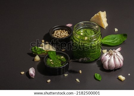 Classic Italian basil pesto sauce in a glass jar. Homemade food, ingredients for a traditional recipe. Hard light, dark shadow, black stone background, copy space