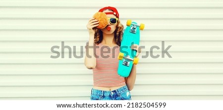 Portrait of stylish young woman with burger and skateboard wearing baseball cap, sunglasses on white background