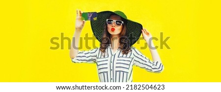 Portrait of beautiful happy young woman taking selfie with smartphone blowing her lips sending air kiss wearing summer straw hat, white striped shirt on yellow background
