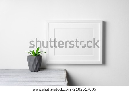 Blank artwork frame mockup over white wall with home plant decor