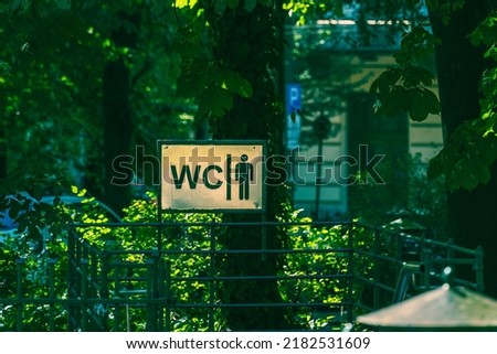 A metal sign indicating a free open-air public toilet in a green area in the shade of leaves on a boulevard in the city of Krakow, a play of sunlight in the shade of trees, a travel service in Poland