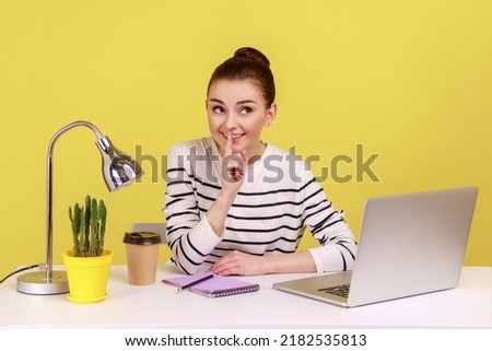 Happy woman employee at workplace shushing with silence gesture, holding finger on lips and smiling to camera, asking to keep secret. Indoor studio studio shot isolated on yellow background.