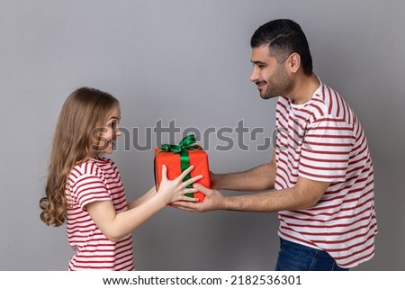 Portrait of satisfied father and daughter in striped T-shirts, man giving present box to his cute child, celebrating birthday together. Indoor studio shot isolated on gray background.