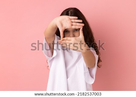Portrait of concentrated little girl wearing white T-shirt looking at camera with one eye, focusing through photo frame made of fingers. Indoor studio shot isolated on pink background.