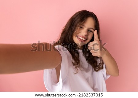 Little girl wearing white T-shirt has optimistic face expression, making selfie, expresses sincere emotions, showing thumb up, point of view photo. Indoor studio shot isolated on pink background.