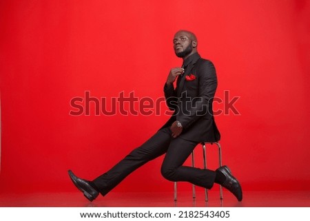 Creative emotional Portraits of a young mid-adult Kenyan black Male Man wearing a black complete suit black tie and red pocket size, black socks, and shoes against a red background in the studio Royalty-Free Stock Photo #2182543405