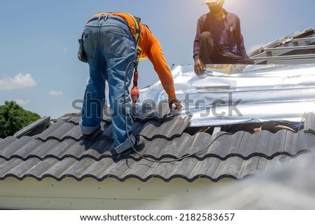 Roof repair, worker replacing gray tiles or shingles on house with blue sky as background and copy space, Roofing - construction worker standing on a roof covering it with tiles. Royalty-Free Stock Photo #2182583657