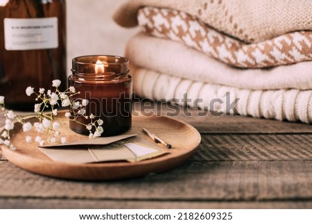 Burning candle in small amber glass jar, flowers of gypsophila and stack knitted seasin sweaters. Cozy lifestyle, hygge concept Royalty-Free Stock Photo #2182609325