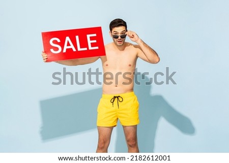 Shirtless handsome Caucasian man wearing summer short pants holding red sale sign in light blue color isolated studio background