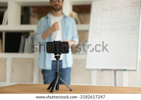 On background man record webinar on cell phone. Tutor business coach stands near flip chart make speech on camera filming educational video for internet audience. Vlogging, tutoring, education concept Royalty-Free Stock Photo #2182617247