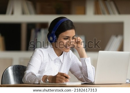 Indian student woman sit at desk do university assignment prepare for exams learn subject use webinar online resources, listen audio course via headphones hold pen noting making task. E-learn concept Royalty-Free Stock Photo #2182617313