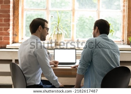 Rear back two colleagues teammates discuss strategy, work on collaborative on-line project sit at desk with laptop, white mock up on device screen. Website, connection, workflow, partnership concept Royalty-Free Stock Photo #2182617487