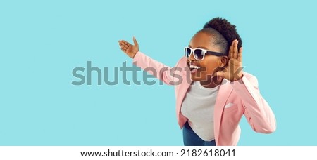 Funny dark-skinned woman puts her hand to her ear as sign that she is listening or eavesdropping. Cheerful woman listening to you pointing with hand on copy space on light blue background. Web banner. Royalty-Free Stock Photo #2182618041