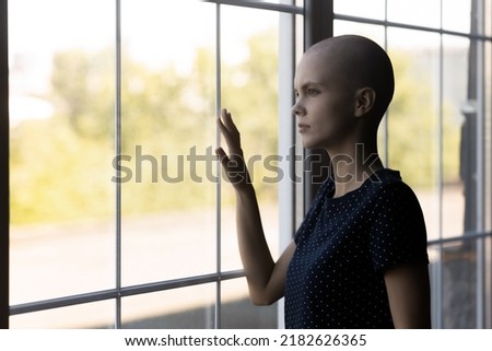 Close up unhappy sick hairless woman lost in thoughts, standing near window, touching glass, looking to aside, thinking about health problem, dreaming about recovery and remission, future Royalty-Free Stock Photo #2182626365