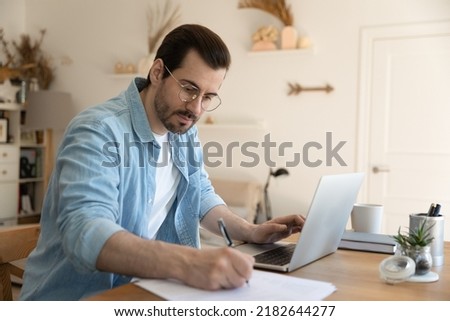 Focused young man in eyeglasses watching educational webinar lecture on computer, writing notes on paper at home office. Serious businessman analyzing electronic marketing documents indoors. Royalty-Free Stock Photo #2182644277