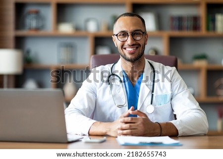Portrait of happy arabic doctor handsome young man in workwear posing at modern clinic, sitting at workdesk with laptop and medical chart, cheerfully smiling at camera, copy space