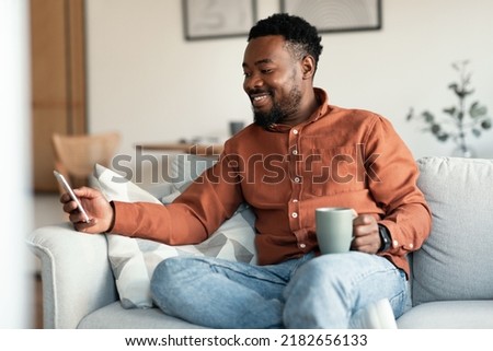 Happy Black Man Using Application On Smartphone And Drinking Coffee Sitting On Sofa At Home. Male Communicating Texting On Cellphone On Weekend. Mobile Communication And Technology