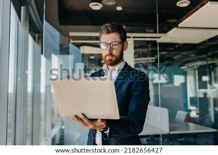 Business person. Successful middle aged businessman with laptop in hands standing in modern office interior. Serious male entrepreneur in formal suit working on computer Royalty-Free Stock Photo #2182656427