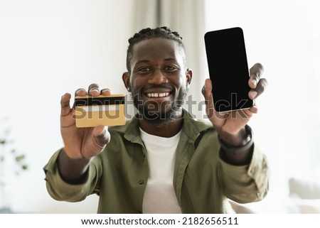 Mobile shopping appliation. Happy black guy holding credit card in hand and showing smartphone with blank screen to camera, free space for mock up, digital pay