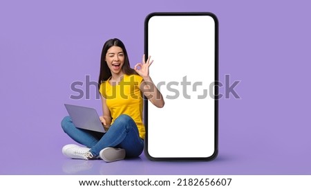 Freelance Jobs, mobile app. Positive young asian lady sitting by huge cell phone with blank screen with laptop on laps and showing ok sign over purple studio background, panorama, copy space, mockup