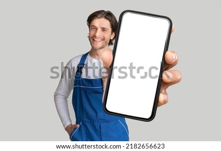 Repairing Service App. Young Handyman In Blue Workwear Showing Modern Mobile Phone With White Blank Screen And Smiling, Posing Over Grey Background With Copy Space, Collage, Mockup Royalty-Free Stock Photo #2182656623