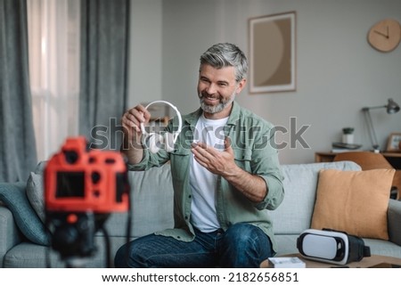 Smiling senior caucasian bearded man blogger records video review on camera, shows headphones, make review in room interior, copy space. Work for retired at home, hobby and overview of devices remote Royalty-Free Stock Photo #2182656851