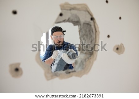 Demolition of an old house. Demolition of unnecessary walls with hand-held impact hammer. A view of an experienced construction worker trying with all might to enlarge hole in wall. Royalty-Free Stock Photo #2182673391
