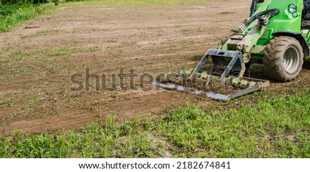 Land work by the territory improvement. Small tractor with a ground leveler for moving soil, turf. A green mini skid steer loader clear the construction site. Machine for agriculture work. Copy space. Royalty-Free Stock Photo #2182674841