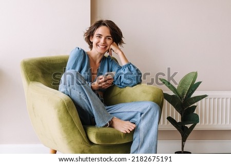 View of beautiful woman in home with phone .Brunette carre hair look and smiling to camera sitting on the green chair at home. Relax, lifestyle concept  Royalty-Free Stock Photo #2182675191