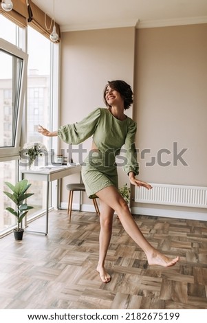 Full-length view of cheerful pretty woman staying in bright room . Caucasian short hair wear green dress smiling and looking at window. Lifestyle, leisure life concept  Royalty-Free Stock Photo #2182675199