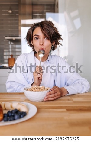 Image of funny woman with spoon at mouth having breakfast . Caucasian beautiful woman sitting on the table with muesli look at camera. Good morning consept 