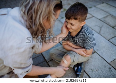 Young mother comforting her little son and giving him bandage after getting hurt when fall down in street. Royalty-Free Stock Photo #2182689211