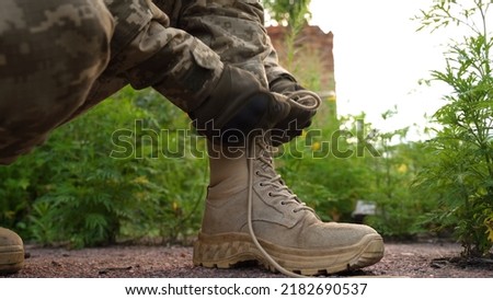 a soldier in a camouflage uniform and tactical gloves laces up his military boots. tactical footwear in combat conditions. preparation for a long-distance combat march. running in combat conditions Royalty-Free Stock Photo #2182690537