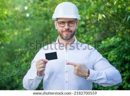 Businessman man in hardhat pointing finger at blank business card outdoors, copy space