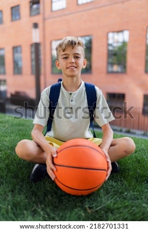 Caucasian boy sitting in public city park with baskaetball ball and looking at camera.