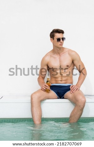 muscular man in swimming trunks and sunglasses sitting at poolside with bottle of beer Royalty-Free Stock Photo #2182707069